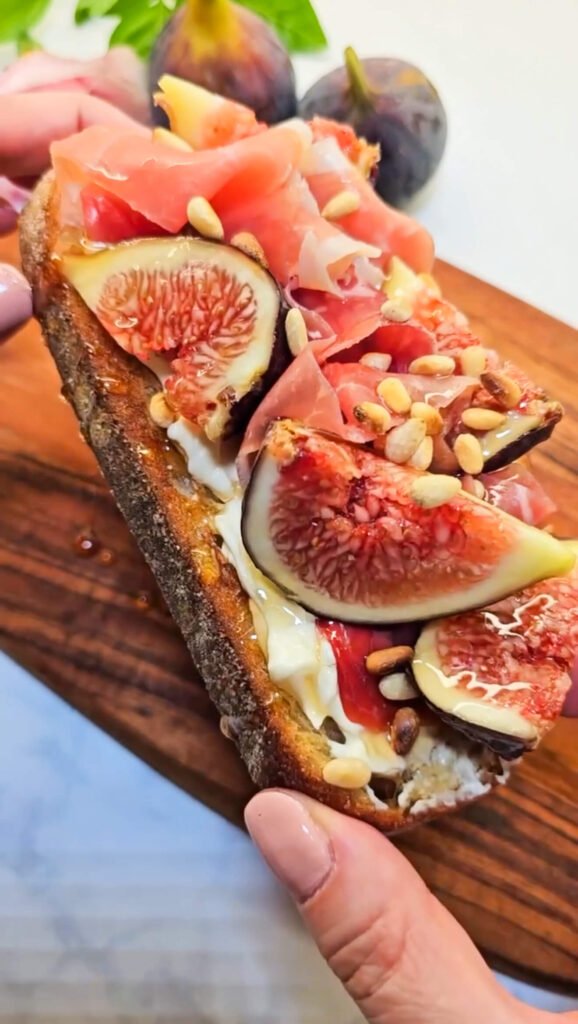 Close-up of a gourmet sourdough toast topped with creamy Brie, fresh fig slices, silky prosciutto, toasted pine nuts, and a drizzle of honey, served on a wooden board with whole figs in the background.