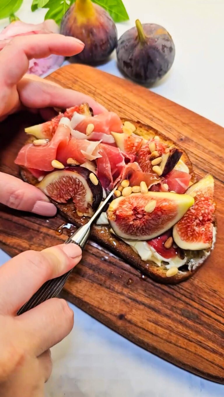 Hands slicing into a fig and prosciutto sourdough toast layered with melted Brie, honey, and toasted pine nuts, on a rustic wooden serving board with fresh figs nearby.