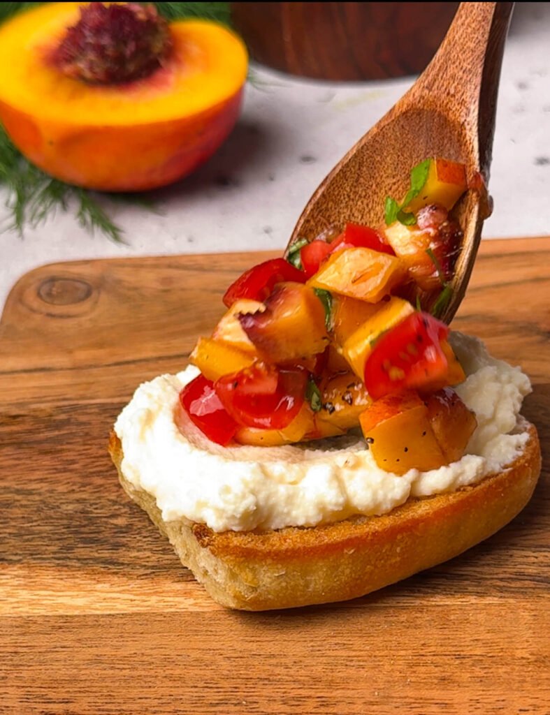 Wooden spoon adding fresh peach and tomato mixture onto ricotta-topped sourdough toast for homemade peach bruschetta