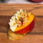 Single peach crostini with piped Cambozola cream cheese, chopped walnuts, and honey drizzle on toasted baguette