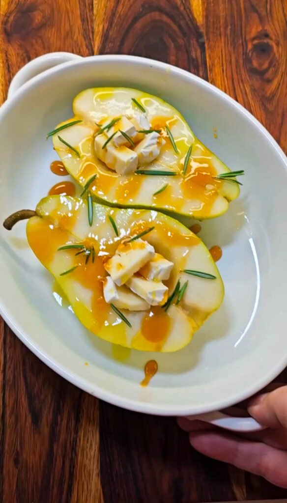 Halved Bartlett pear filled with Brie cheese, drizzled with honey, lime juice, and rosemary leaves in a white baking dish