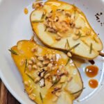 Oven-baked pear halves topped with melted Brie, toasted pine nuts, honey drizzle, and rosemary in a white serving dish