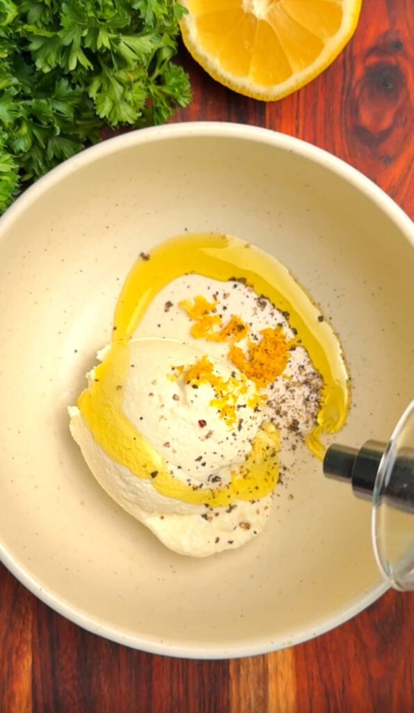 Bowl of ricotta cheese mixed with lemon zest, cracked black pepper, and a drizzle of olive oil, with fresh parsley and half a lemon beside it on a wood surface.