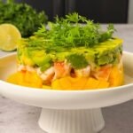 A plated shrimp and mango stack layered with diced mango on the bottom, chopped shrimp in the middle, and avocado on top, garnished with fresh microgreens and drizzled dressing.