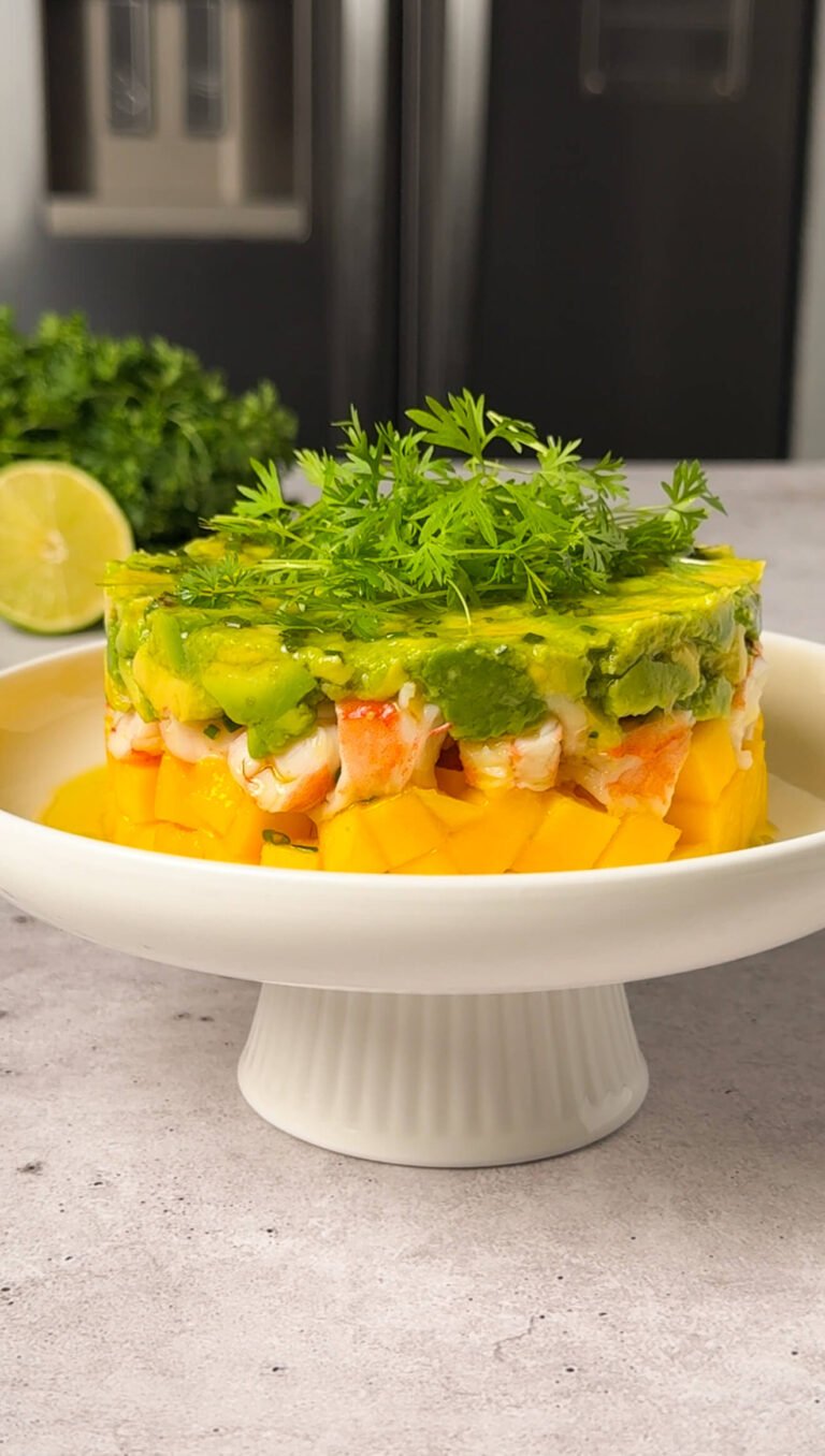 A plated shrimp and mango stack layered with diced mango on the bottom, chopped shrimp in the middle, and avocado on top, garnished with fresh microgreens and drizzled dressing.