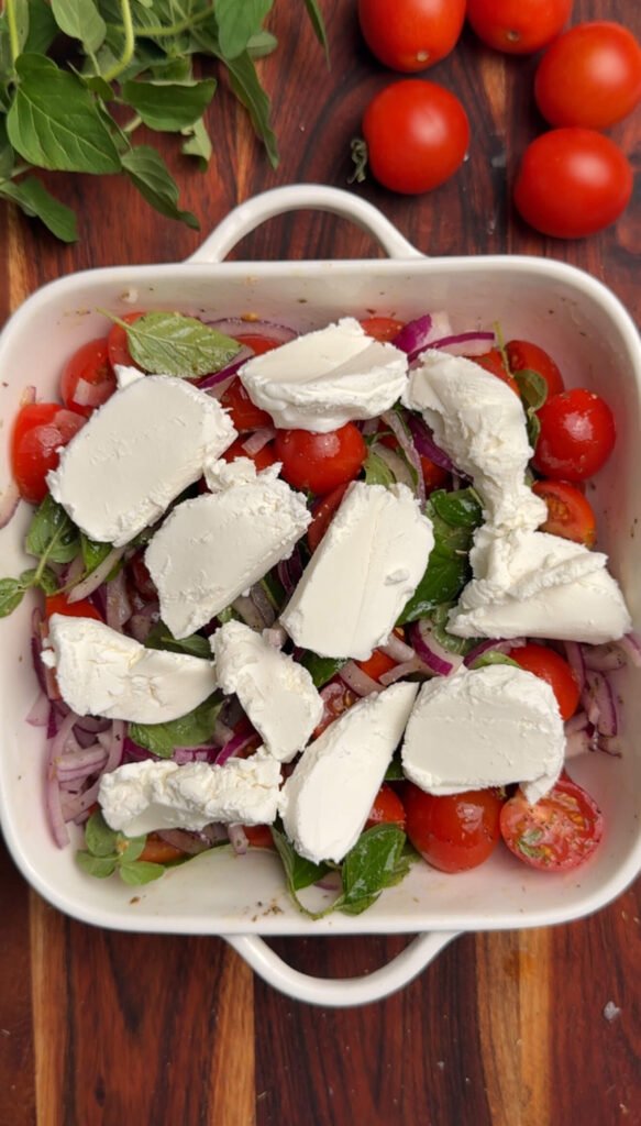 lices of creamy goat cheese arranged on top of seasoned cherry tomatoes, red onion, and oregano in a baking dish before baking.