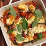 Golden, bubbly tomato bake with goat cheese in a white baking dish, garnished with fresh oregano leaves.