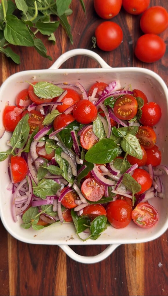 Cherry tomatoes, sliced red onion, and fresh oregano tossed together in a white baking dish with olive oil and seasoning.