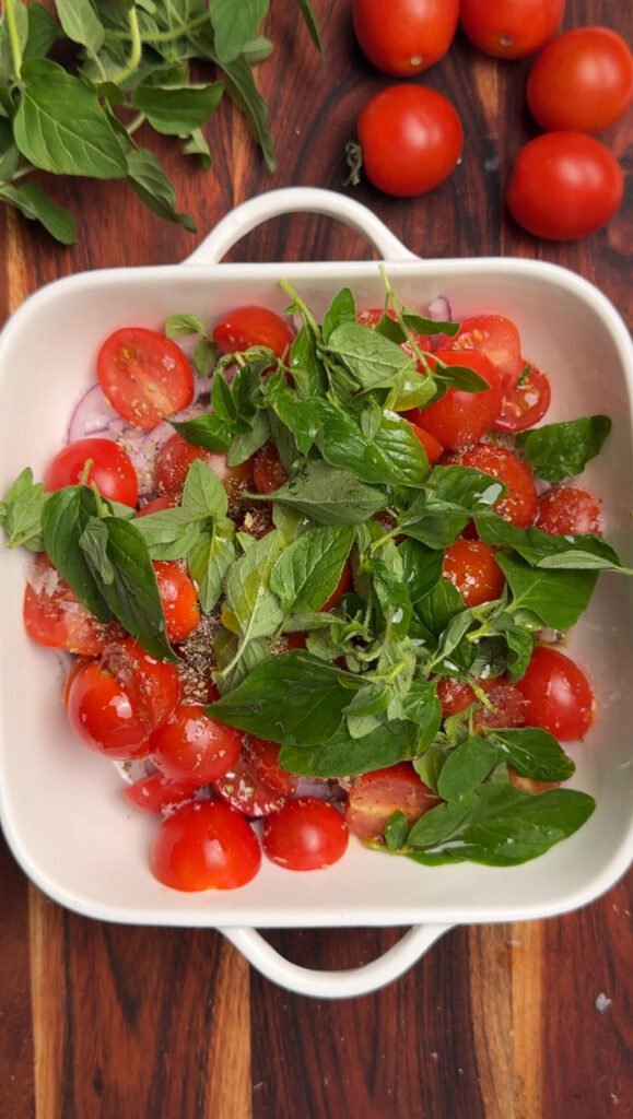 Overhead view of a white baking dish filled with halved cherry tomatoes, fresh oregano leaves, and seasonings, ready for roasting.