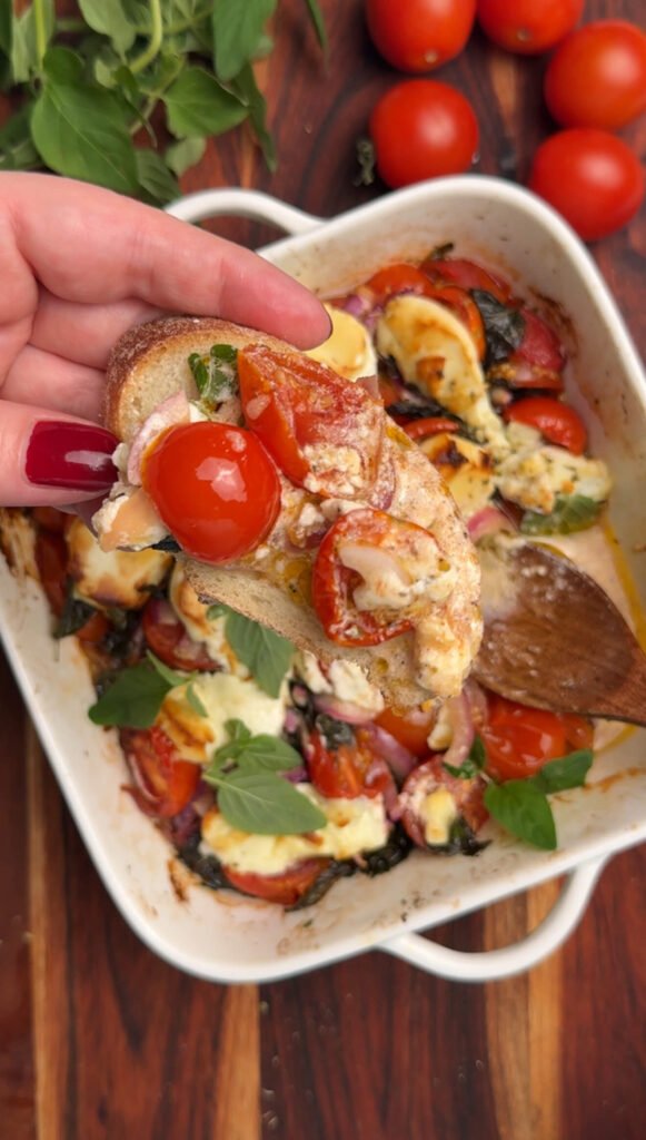 Close-up of a toasted bread slice topped with roasted cherry tomatoes, melted goat cheese, and fresh oregano leaves.