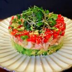 A beautifully layered Spicy Shrimp Stack on a white plate, featuring avocado, shrimp, fresh tomato-cucumber salsa, sesame seeds, and microgreens on top.