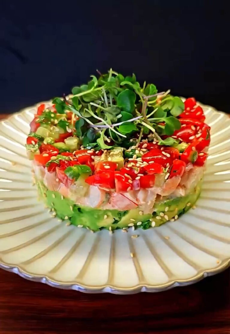 A beautifully layered Spicy Shrimp Stack on a white plate, featuring avocado, shrimp, fresh tomato-cucumber salsa, sesame seeds, and microgreens on top.