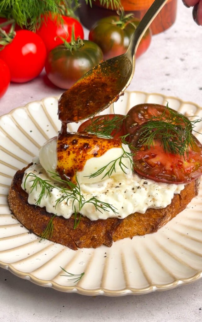 A slice of sourdough toast layered with cottage cheese, sliced tomatoes, and a poached egg, while a spoon drizzles smoky Aleppo pepper butter over the egg. Fresh dill sprigs finish the dish, making it look bold, rustic, and flavorful.