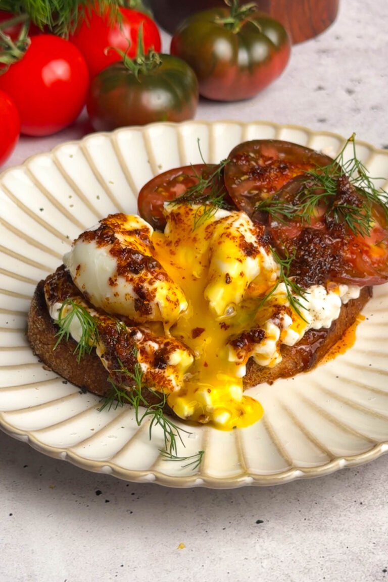 A golden sourdough toast topped with cottage cheese, sliced tomatoes, and a poached egg, cut open to reveal a rich, runny yolk. Warm Aleppo pepper butter drizzles over the toast, garnished with fresh dill, creating a vibrant and indulgent presentation.