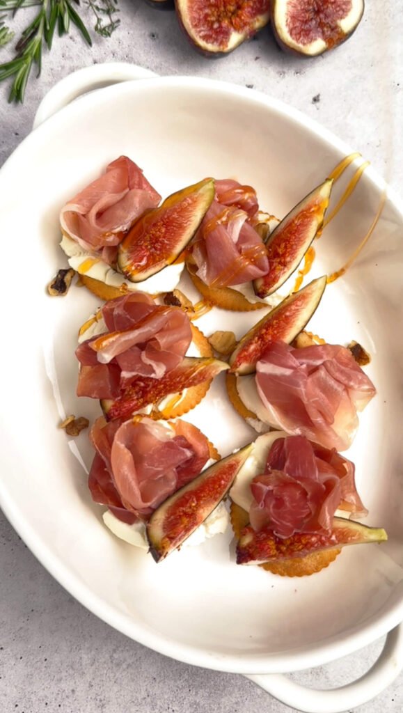 Freshly baked goat cheese bites topped with prosciutto rosettes and fig slices, drizzled with hot honey in a white baking dish — warm charcuterie-style appetizer.