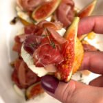 Hand holding one baked cheese bite with goat cheese, ripe fig slice, and prosciutto rosette over a white dish — creamy, salty, and sweet bite-sized appetizer.