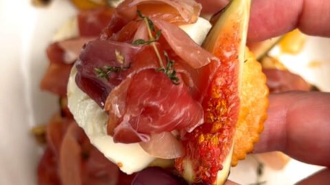 Hand holding one baked cheese bite with goat cheese, ripe fig slice, and prosciutto rosette over a white dish — creamy, salty, and sweet bite-sized appetizer.