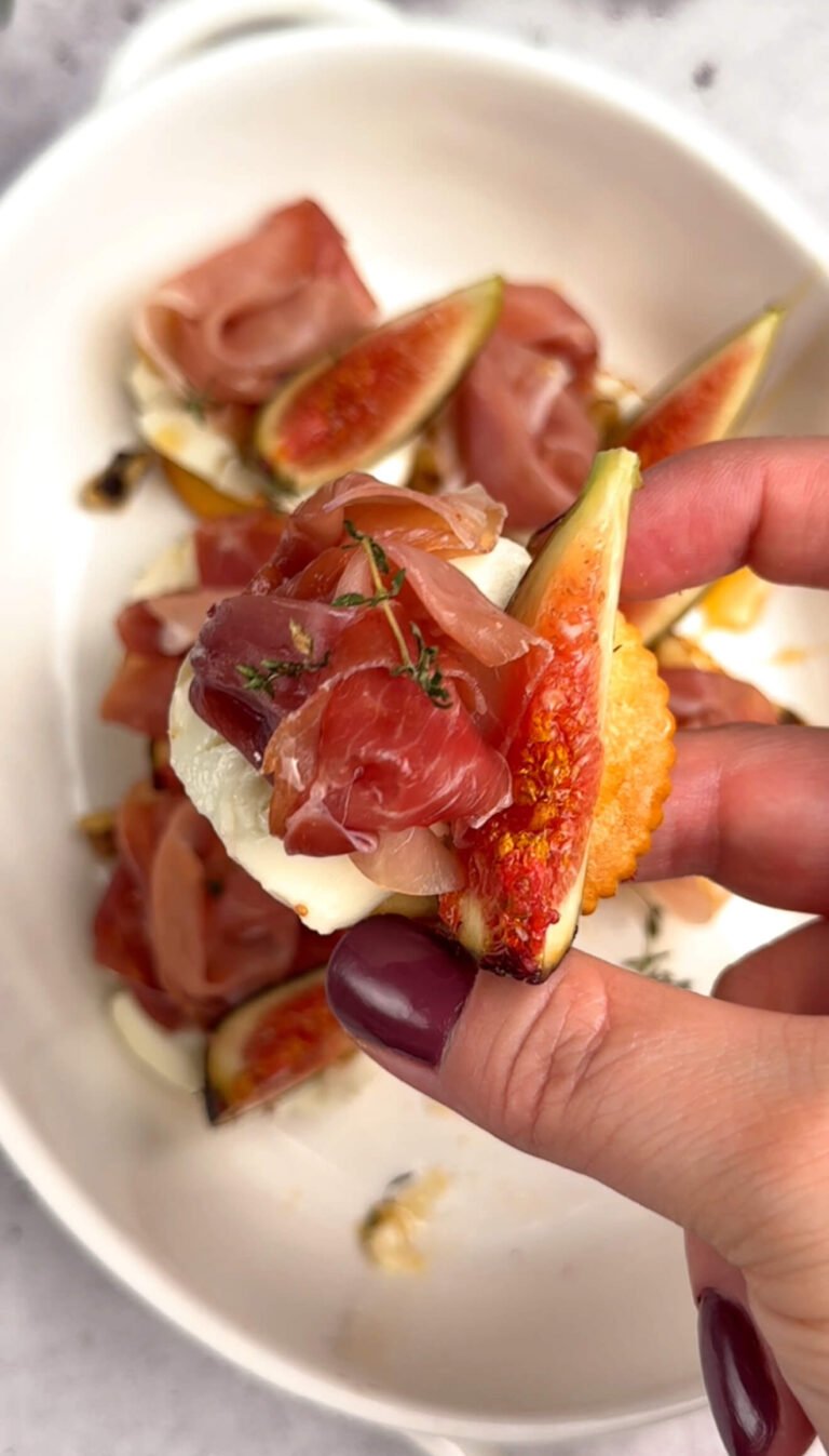 Hand holding one baked cheese bite with goat cheese, ripe fig slice, and prosciutto rosette over a white dish — creamy, salty, and sweet bite-sized appetizer.
