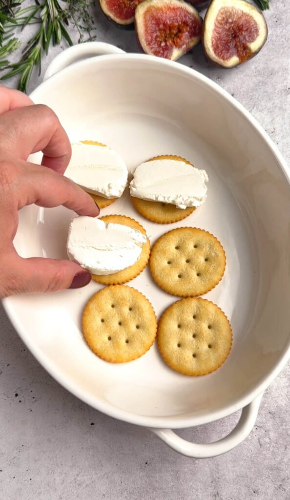 Step-by-step photo showing Ritz crackers topped with slices of goat cheese before baking — beginning stage of baked cheese bites with figs and prosciutto.