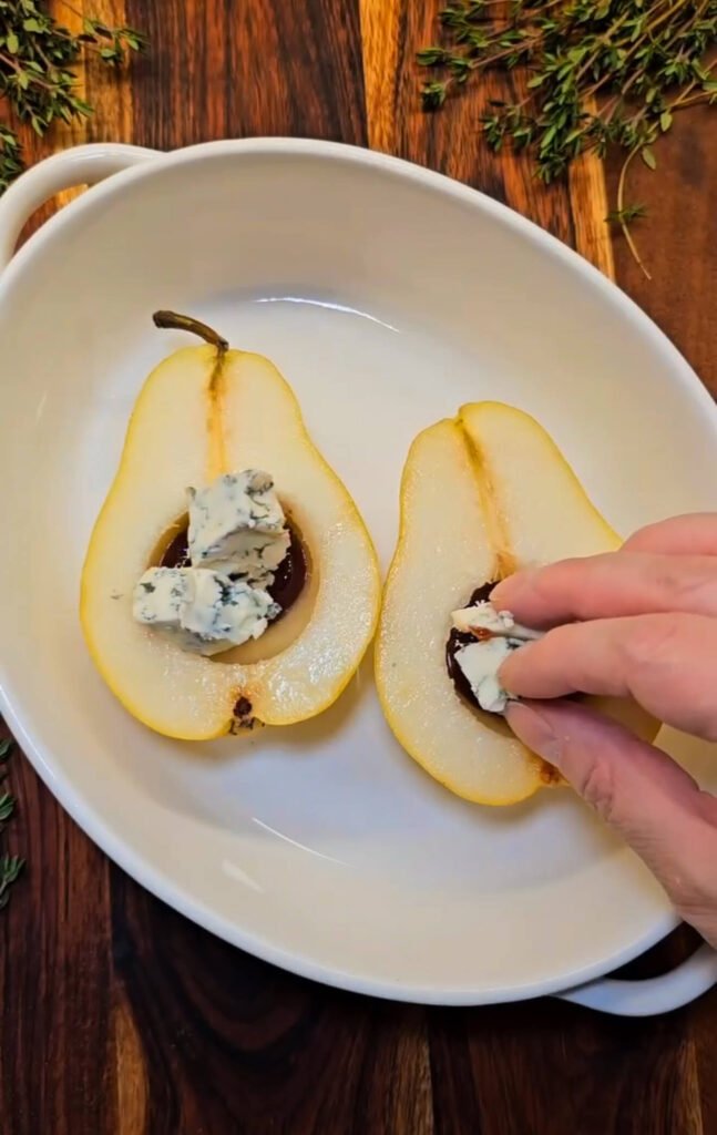 Bartlett pear halves filled with plum jam and chunks of Gorgonzola cheese before baking, surrounded by fresh thyme on a wooden board