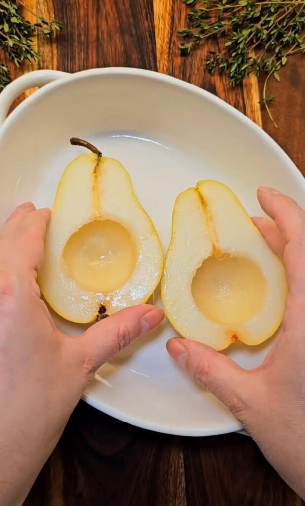 Two fresh Bartlett pear halves with centers scooped out, arranged in a white baking dish on a wooden surface with sprigs of thyme