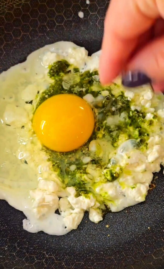 Feta cheese melting in a skillet with a spoonful of basil pesto and a cracked egg in the center, sizzling together to create a rich, savory base for toast.