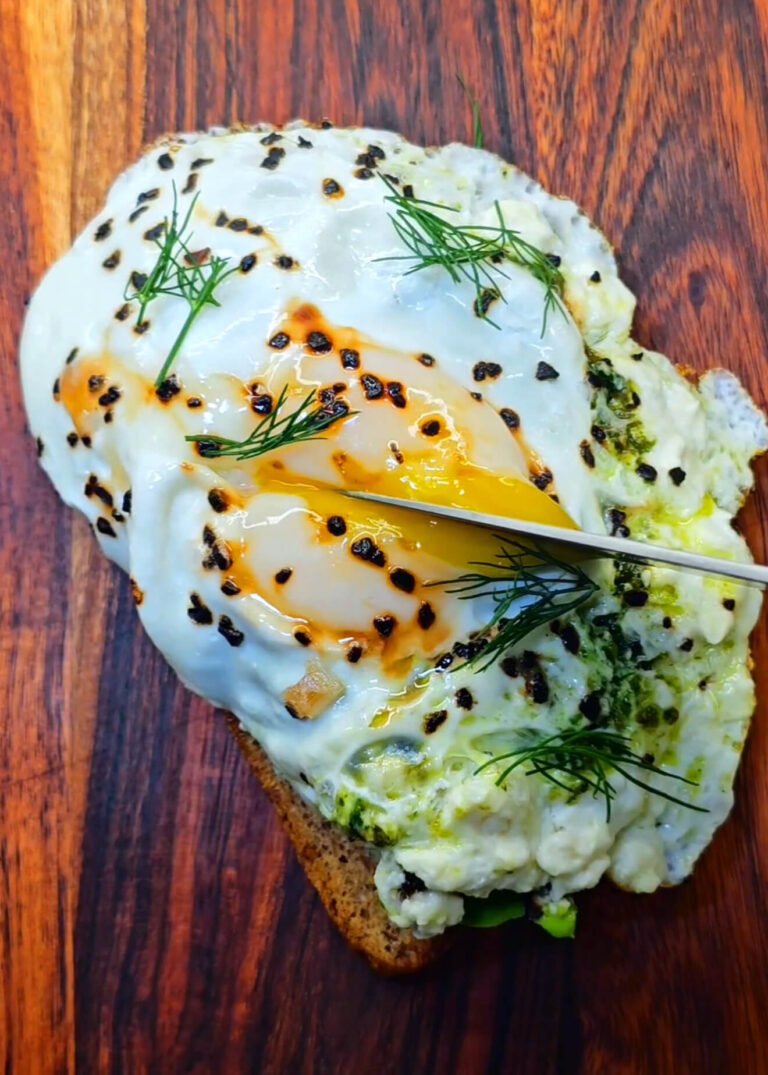 Golden fried egg with melted feta and pesto layered on toasted bread, sprinkled with granulated black garlic and topped with fresh dill, ready to be sliced open.