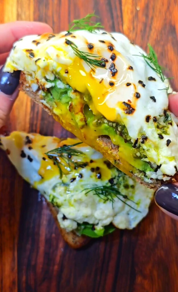Close-up of cozy breakfast toast with feta, pesto, and fried egg seasoned with black garlic. The golden yolk drips over smashed avocado and melted feta, garnished with fresh dill on rustic bread.