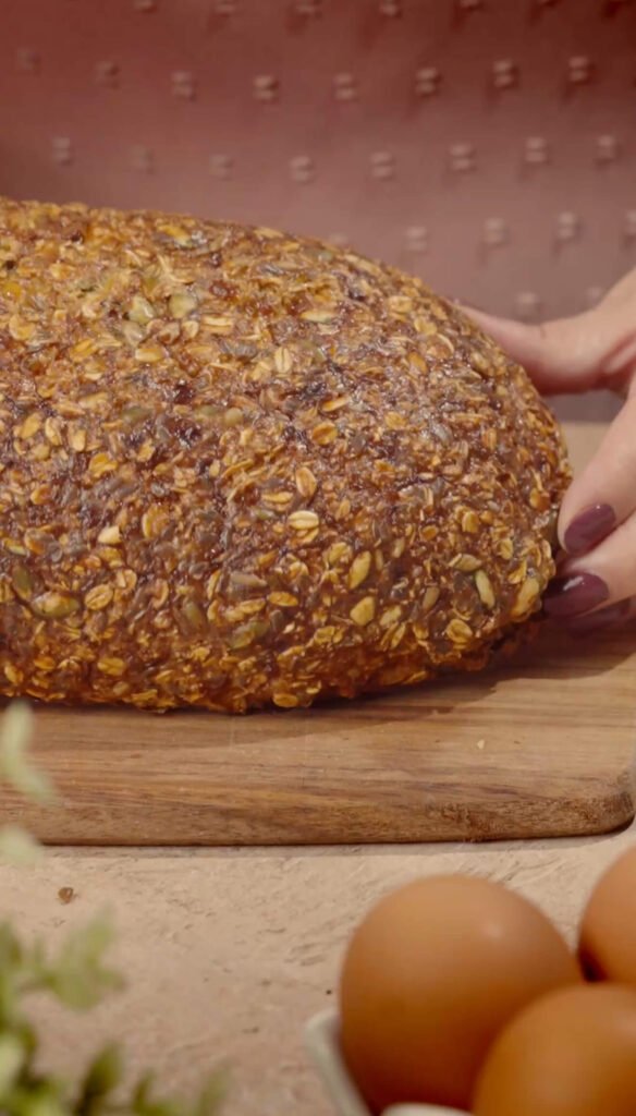 A golden-brown, rustic loaf of gluten-free fitness bread covered with oats and seeds resting on a wooden board, with a hand gently holding the side.