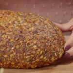 A golden-brown, rustic loaf of gluten-free fitness bread covered with oats and seeds resting on a wooden board, with a hand gently holding the side.