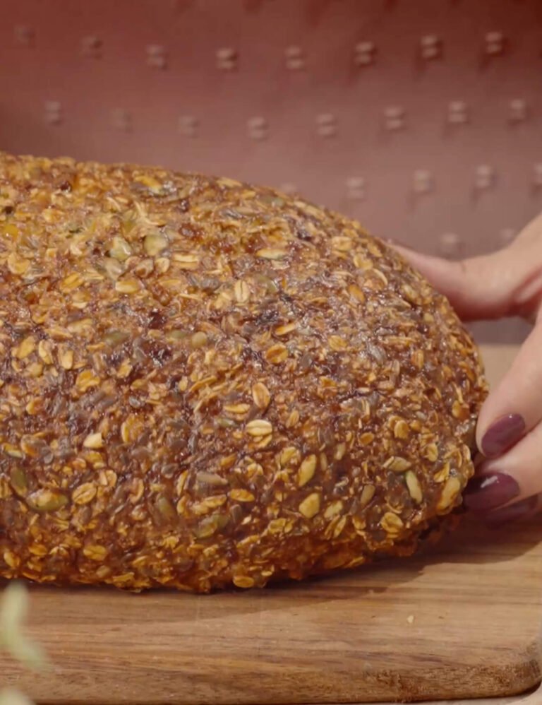 A golden-brown, rustic loaf of gluten-free fitness bread covered with oats and seeds resting on a wooden board, with a hand gently holding the side.