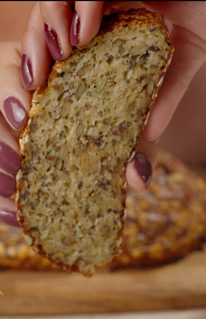 A close-up of a slice of gluten-free seed bread held between two hands, showing its moist, hearty texture filled with oats and seeds.