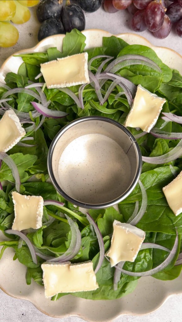 Arugula and spinach arranged in a wreath shape on a cream platter with thinly sliced red onion and pieces of Brie cheese evenly spaced around the greens.