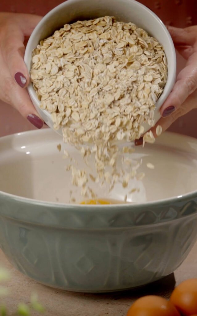 Rolled oats being poured from a white bowl into a mixing bowl with eggs at the bottom, part of the preparation for homemade gluten-free fitness bread.