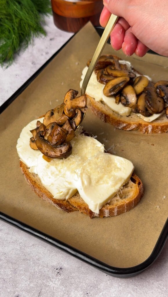 Mozzarella-covered sourdough being topped with freshly cooked mushrooms using a golden spoon, ready to be baked into cheesy mushroom toast. The image shows the layering process with Parmesan and earthy mushrooms glistening with balsamic glaze.