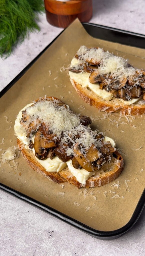 Two slices of sourdough toast layered with mozzarella, Parmesan, and sautéed mushrooms on a baking sheet lined with parchment paper — ready to be baked until the cheese is golden and bubbly.