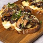 Finished double cheese mushroom toasts on a wooden board, topped with fresh thyme and melted mozzarella bubbling beneath caramelized mushrooms. The scene evokes a warm, rustic, and cozy fall feeling