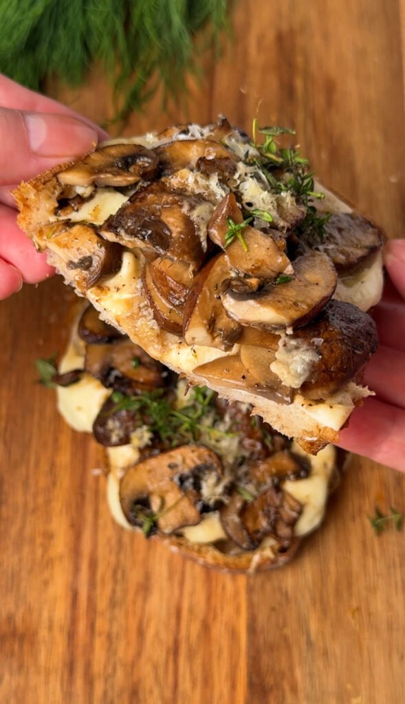 A close-up of a hand holding a slice of double cheese mushroom toast topped with golden sautéed mushrooms, melted mozzarella, and fresh thyme on toasted sourdough. The gooey cheese and glistening mushrooms highlight the cozy, savory texture of the toast.