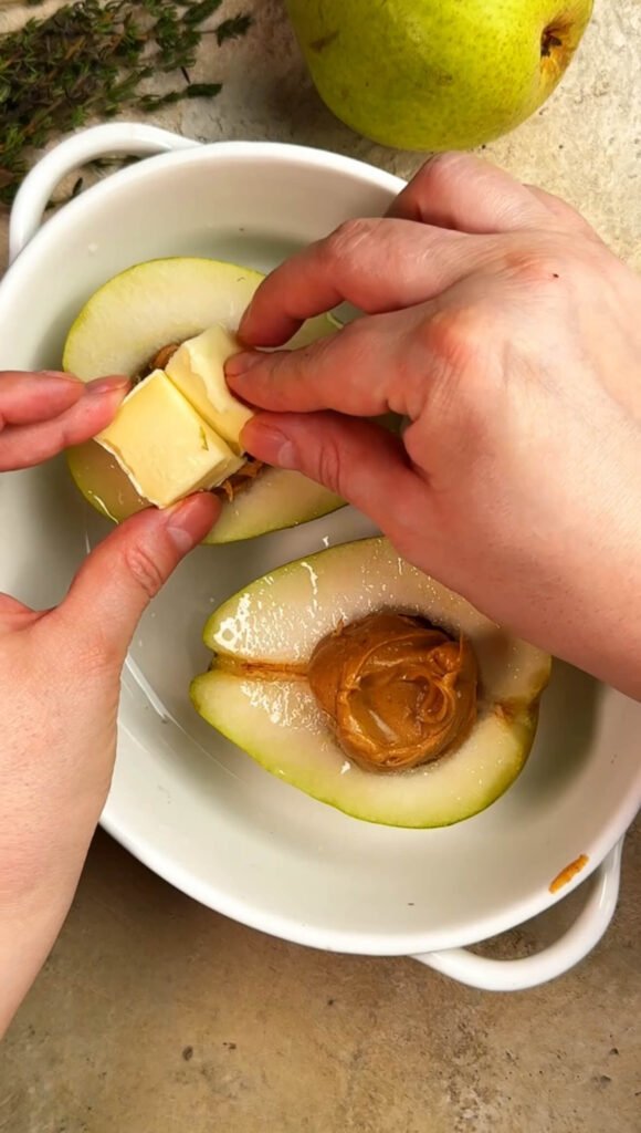Hands adding cubes of creamy brie onto halved pears filled with smooth peanut butter before baking