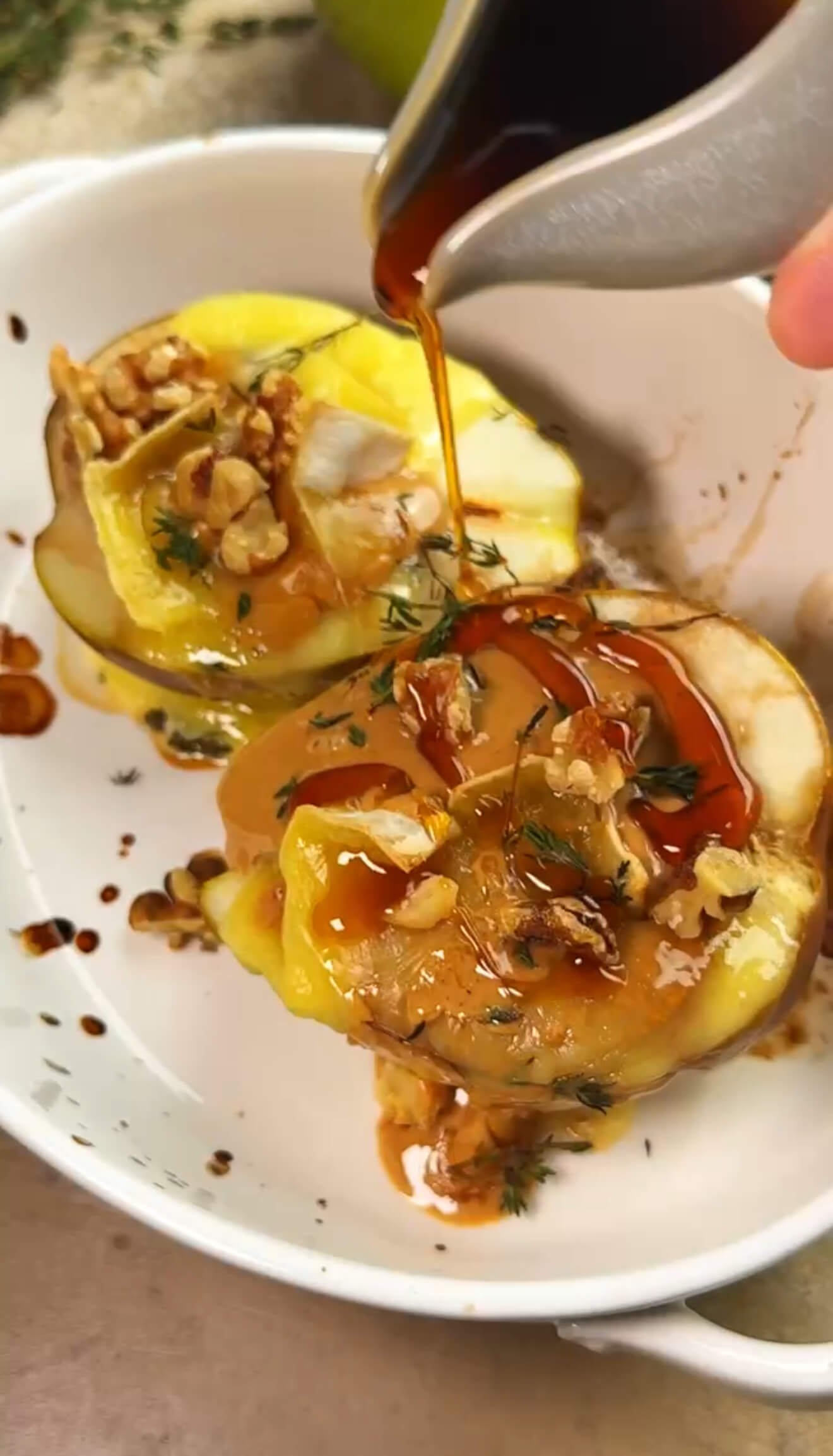 Warm baked pear topped with melted brie, creamy peanut butter, toasted walnuts, and fresh thyme as maple syrup is being poured over