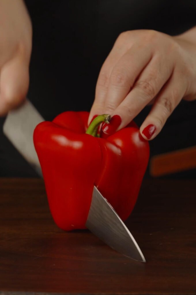 Red bell pepper being sliced on a cutting board — the vibrant color adds freshness and sweetness to the tomato sauce base for the shakshuka.