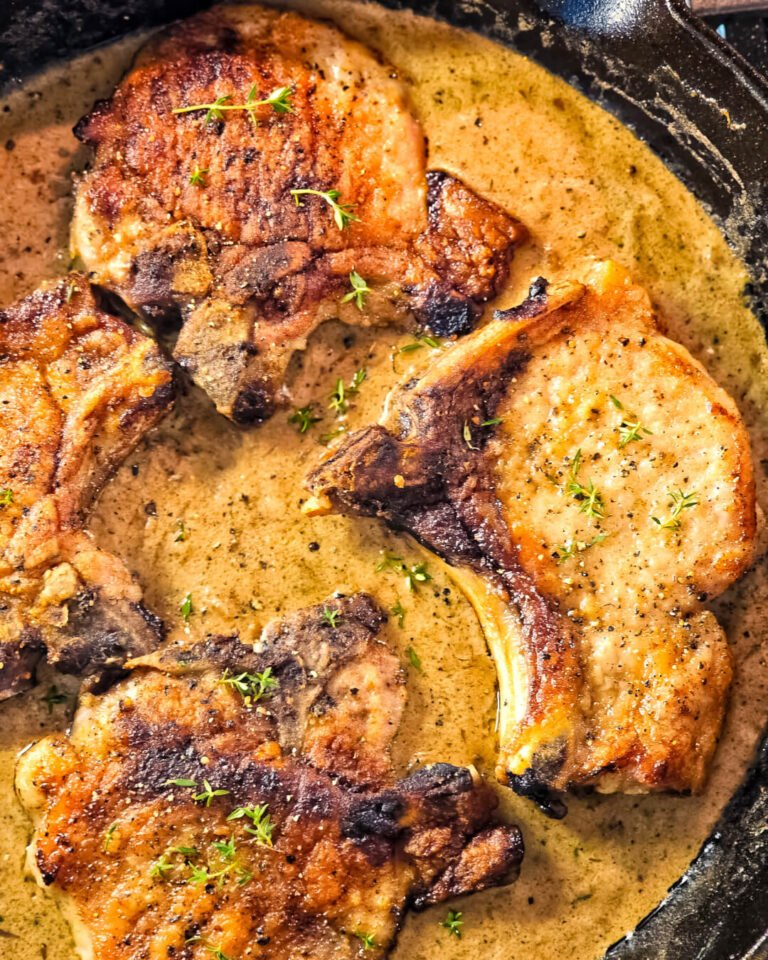 Golden-seared bone-in pork chops simmering in a rich, creamy peppercorn sauce inside a cast-iron skillet, garnished with fresh thyme. The sauce has a velvety texture with visible specks of black pepper, highlighting the cozy, rustic feel of this comforting weeknight dinner.