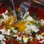 Portobello Shakshuka served in a skillet — a fork cutting into the mushroom cap as the golden egg yolk flows into the tomato sauce, topped with feta and fresh herbs.