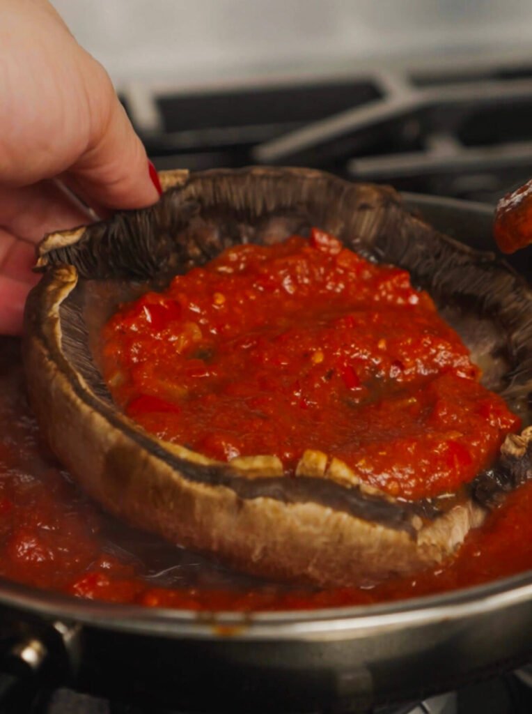 A hand gently placing tomato sauce inside a Portobello mushroom cap over a skillet filled with simmering spiced tomato shakshuka sauce.