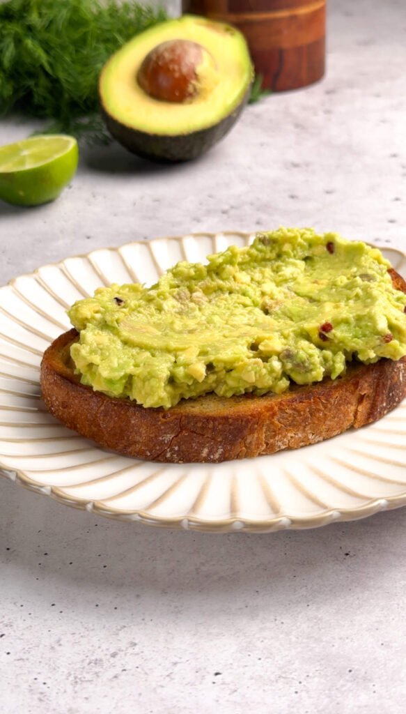 Close-up of mashed avocado spread evenly on a toasted slice of sourdough bread, showing its creamy texture with bits of red pepper flakes and lime. A halved avocado and lime rest in the background.