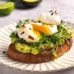 A slice of golden sourdough toast topped with mashed avocado, fresh microgreens, and two perfectly poached eggs with runny yolks, sprinkled with red pepper flakes and drizzled with olive oil. Half an avocado and lime sit blurred in the background on a light gray surface.