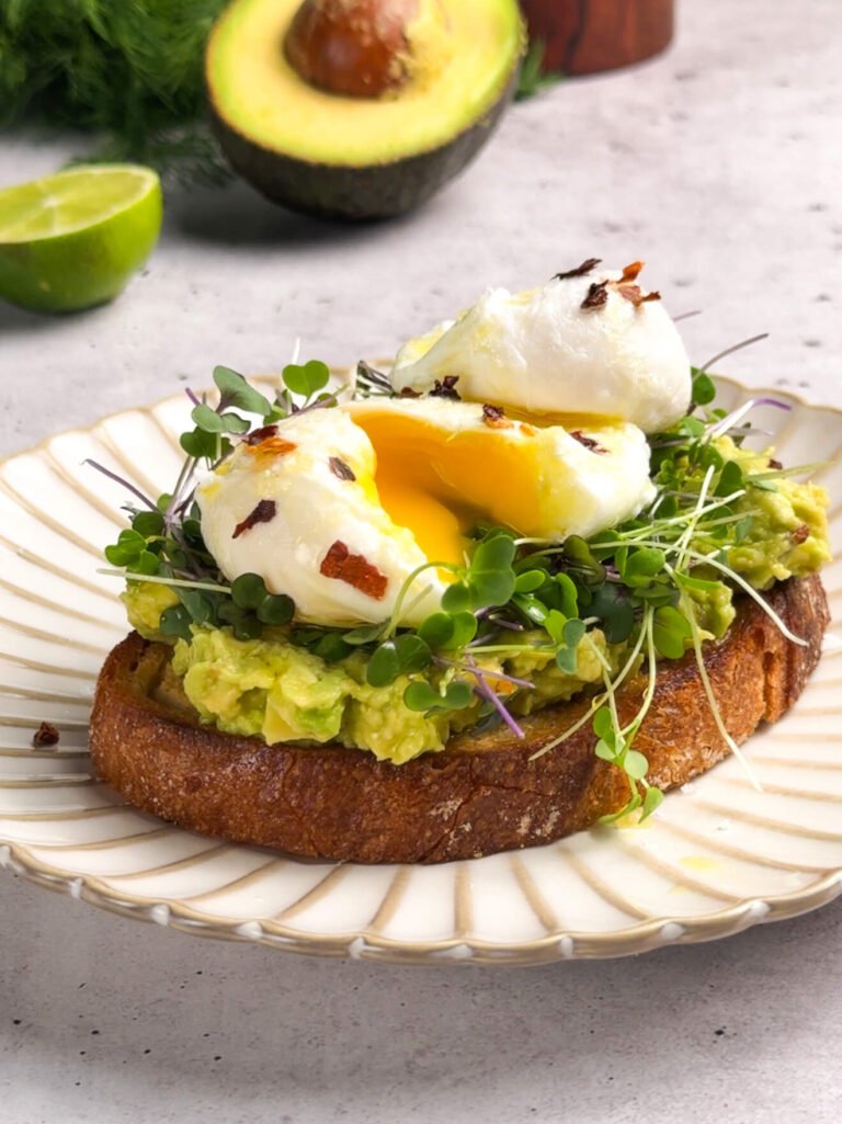 A slice of golden sourdough toast topped with mashed avocado, fresh microgreens, and two perfectly poached eggs with runny yolks, sprinkled with red pepper flakes and drizzled with olive oil. Half an avocado and lime sit blurred in the background on a light gray surface.