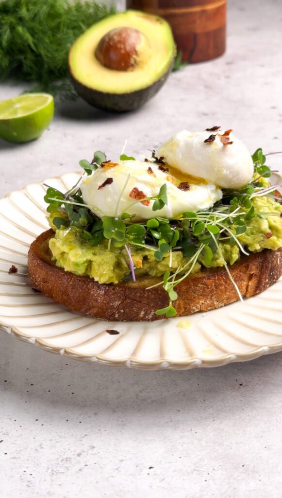 Avocado toast topped with microgreens and two poached eggs glistening with olive oil and sprinkled with red pepper flakes, styled on a scalloped white plate with avocado and lime in the background.