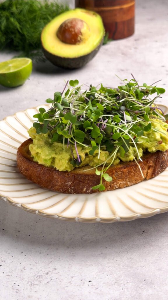 Sourdough toast layered with mashed avocado and topped with a generous handful of fresh green microgreens. Bright avocado halves and lime slices create a fresh, inviting backdrop.