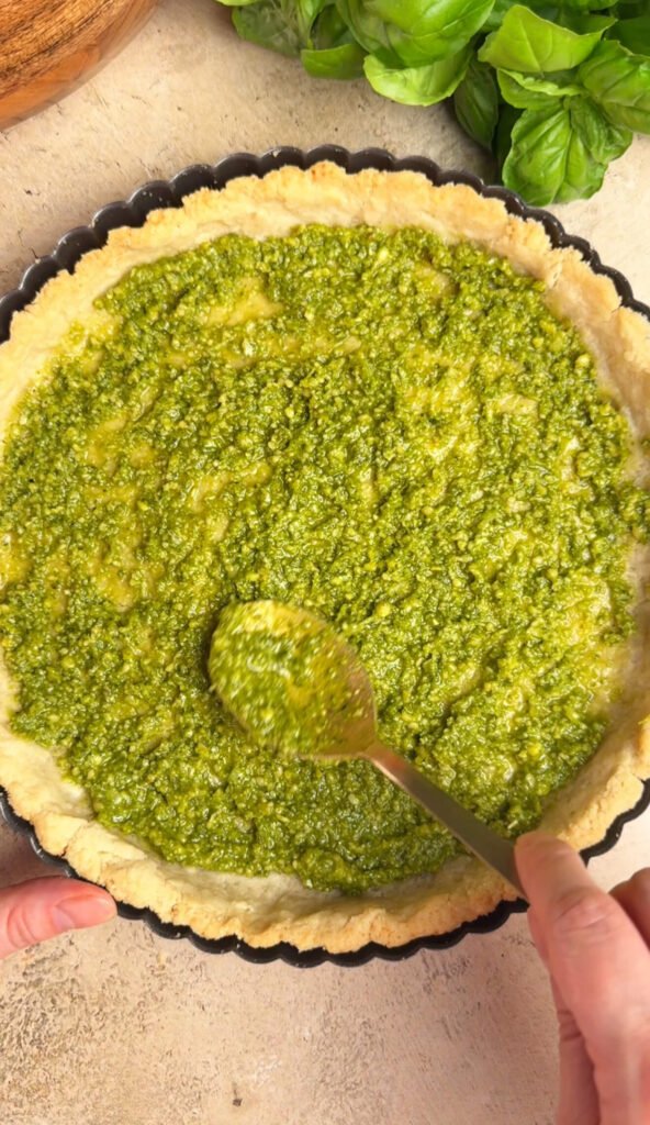 Spreading fresh basil pesto over pre-baked tart crust as the first layer for heirloom tomato pie with ricotta and bacon